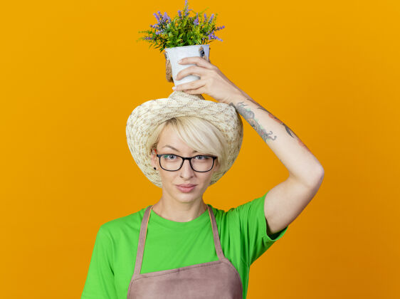 女人一位年轻的园丁 围裙上留着短发 头上戴着帽子 头上抱着盆栽植物 站在橙色背景下 看着相机 自信地微笑着抱着微笑植物