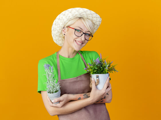 头发一位年轻的园丁 围裙上留着短发 戴着帽子 手里拿着盆栽植物 开心地看着站在橙色背景上的摄像机快乐女人橙色