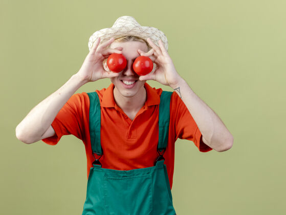 戴着年轻的园丁 穿着连体衣 戴着帽子 手里拿着新鲜的西红柿 站在明亮的背景下微笑着遮住眼睛年轻微笑眼睛