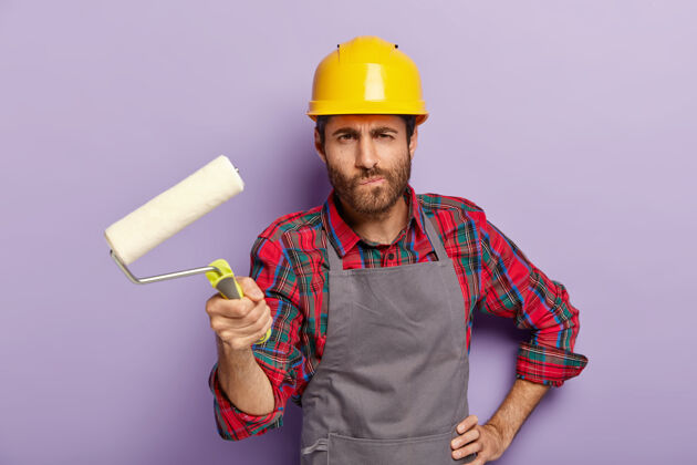 站立严肃的男油漆工拿着油漆辊 在家里重新装修 粉刷墙壁 戴着防护帽和围裙 在室内摆姿势 忙着修理和翻新 孤立在紫色的墙上翻新工匠翻新