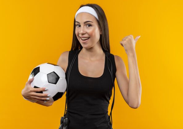 漂亮快乐的年轻漂亮的运动女孩戴着头带和腕带拿着足球 指着一边 脖子上系着跳绳孤立在橙色的空间里指着球女孩