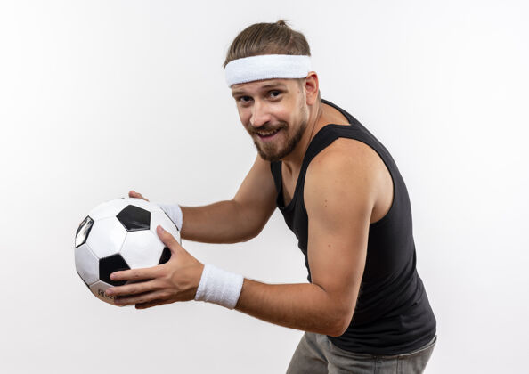 英俊面带微笑的年轻帅气的运动型男子戴着头带和腕带站在侧视图中 拿着足球隔离在空白处足球持有立场