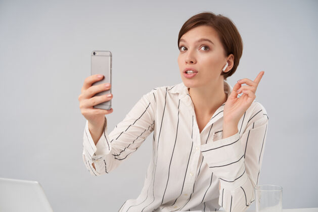 年轻年轻的棕色眼睛短发黑发女商人坐在白色条纹正式衬衫上 用智能手机打视频电话的画像困惑肖像姿势