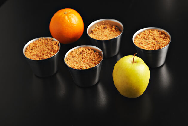 皮一套四个相同的不锈钢杯与苹果碎甜点 一个橙色和一个黄色的苹果拍摄从顶部在黑色的表 侧视图个人的平底锅餐
