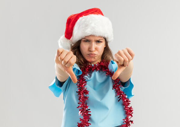 顶戴着蓝色上衣和圣诞帽的年轻女子 脖子上戴着金属丝 看起来很不高兴地伸出大拇指女人圣诞站