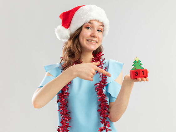 脖子一位身穿蓝色上衣 头戴圣诞帽 脖子上戴着金属箔的年轻女子 手里拿着新年约会的玩具立方体 用食指指着立方体 快乐而快乐年轻玩具指着