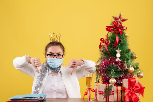 女医生正面图：女医生戴着无菌口罩坐在黄色桌子上 桌上放着圣诞树和礼品盒坐着圣诞节男人