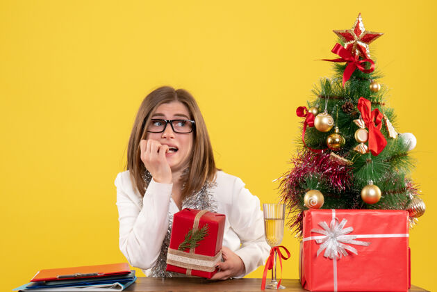 电脑正面图：女医生坐在桌子前 拿着礼物 黄色背景上的圣诞树和礼盒人健康树