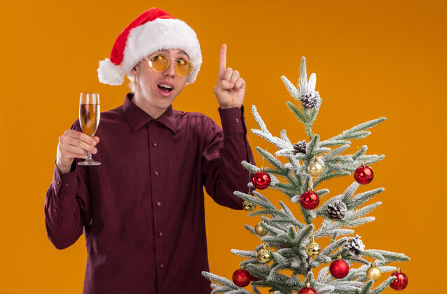 男人令人印象深刻的年轻金发男子戴着圣诞帽和眼镜站在装饰圣诞树附近拿着香槟酒杯看着一边指着孤立的橙色背景侧着杯子圣诞树