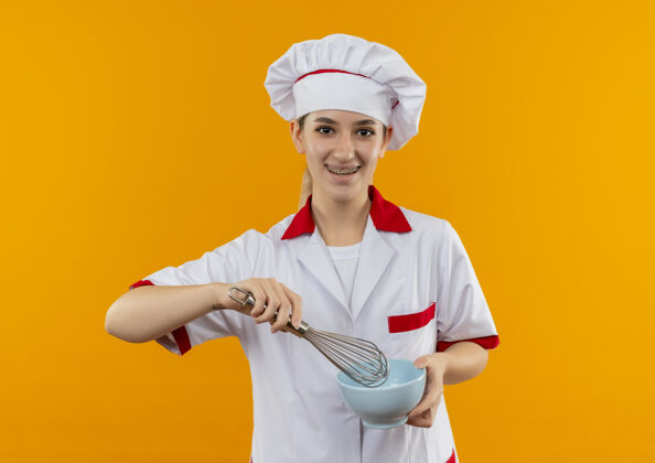 拂快乐的年轻漂亮厨师 身着厨师制服 戴着牙套 拿着搅拌器和碗 隔离在橙色的空间里碗快乐牙套
