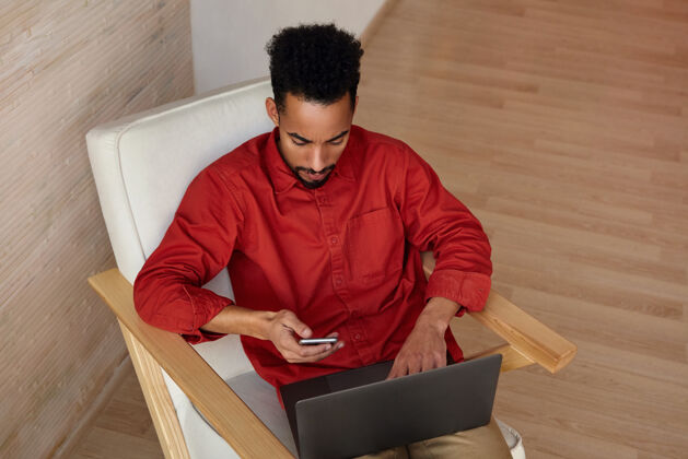 短款一幅年轻的黑发黑皮肤自由职业者的横向肖像 穿着红衬衫 拿着笔记本电脑和智能手机在家里工作 坐在米色内饰的椅子上室内男性专注