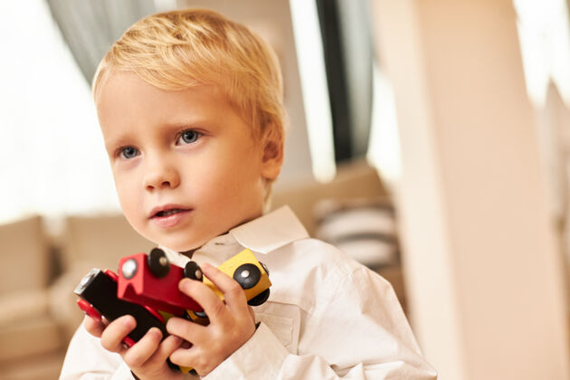 活动帅气的金发欧洲男孩的肖像摆在时尚的客厅室内穿着白衬衫享受室内游戏玩五颜六色的货车或汽车创造力 想象力和幻想学习玩具游戏