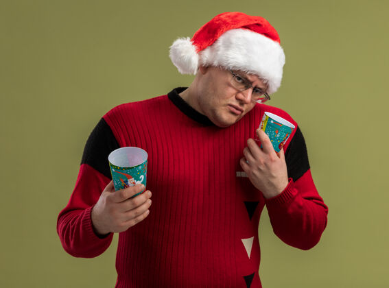 圣诞老人困惑的成年男子戴着眼镜 戴着圣诞帽 举着圣诞咖啡杯 头对侧地看着孤立在橄榄绿背景上的相机圣诞帽子倾斜