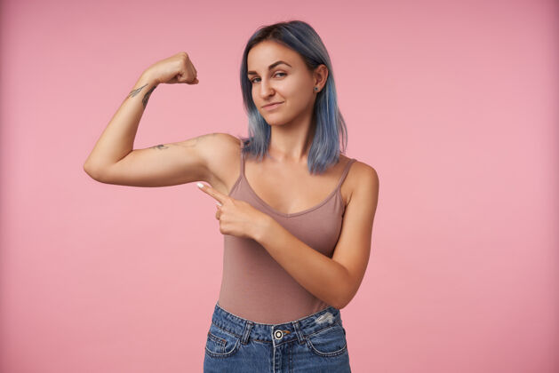 灰色年轻的纹身女性的肖像 短发指向她举起的手 同时显示她强壮的二头肌 站在粉红色的穿着纹身休闲
