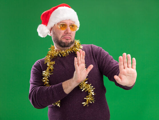 脖子一个戴着圣诞老人帽 脖子上戴着金箔花环 戴着眼镜的中年男人 在绿色背景下 看着一边做着拒绝的手势拒绝花环未租
