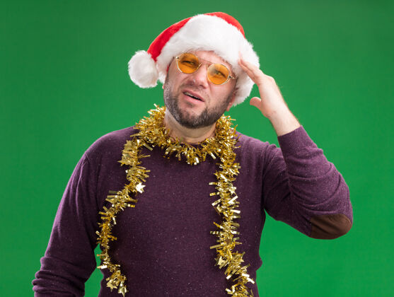 眼镜困惑的中年男子戴着圣诞老人帽 脖子上戴着金属丝花环 戴着眼镜 看着侧碰的头 隔离在绿色背景上男人侧面脖子
