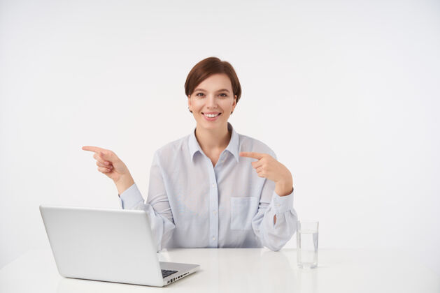 休闲快乐的年轻漂亮的短发黑发女人在办公室里拿着她的笔记本电脑 面带微笑 正眼看着 坐在白色的椅子上用食指指着旁边现代员工女人
