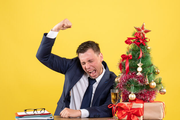 手势一个英俊的男人坐在圣诞树旁的桌子前展示胜利的姿态 黄色的礼物坐着赢公司