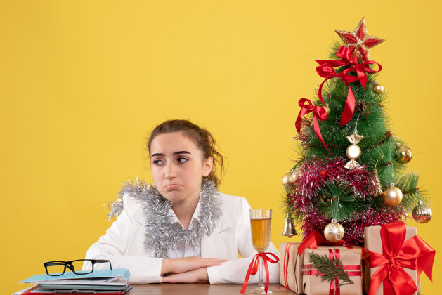 背景正面图：女医生坐在黄色背景的桌子后面 还有圣诞树和礼品盒情感圣诞节快乐