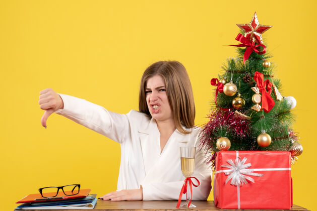 背景前视图：坐在桌子前的女医生不高兴地坐在黄色背景上 上面放着圣诞树和礼品盒人不高兴笔记本电脑