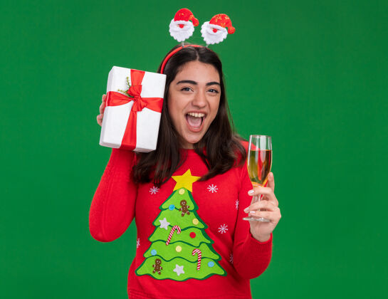 新的快乐的年轻白人女孩戴着圣诞老人的头带 手拿一杯香槟和圣诞礼盒 在绿色背景上与复制空间隔离开来快乐快乐盒子