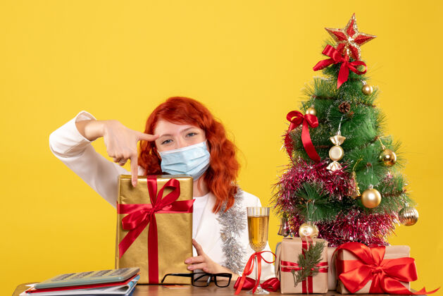 坐着正面图：戴着面具坐着的女医生带着圣诞礼物 黄色背景上的圣诞树和礼盒树快乐成人