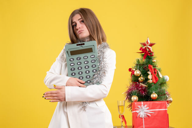 乐器正面图女医生站着拿着计算器 背景是黄色的圣诞树和礼品盒乐器圣诞节护士