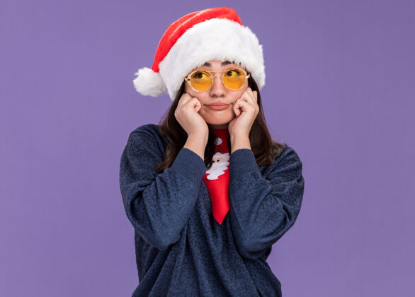领带戴着太阳眼镜 戴着圣诞帽 系着圣诞领带 心烦意乱的白人女孩把手放在脸上 看着侧面太阳年侧