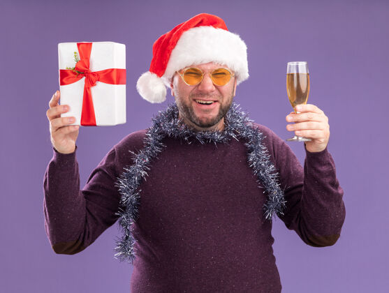 穿着快乐的中年男子戴着圣诞帽 脖子上戴着金箔花环 戴着眼镜 手里拿着礼包 紫色的墙上隔着一杯香槟圣诞老人香槟周围