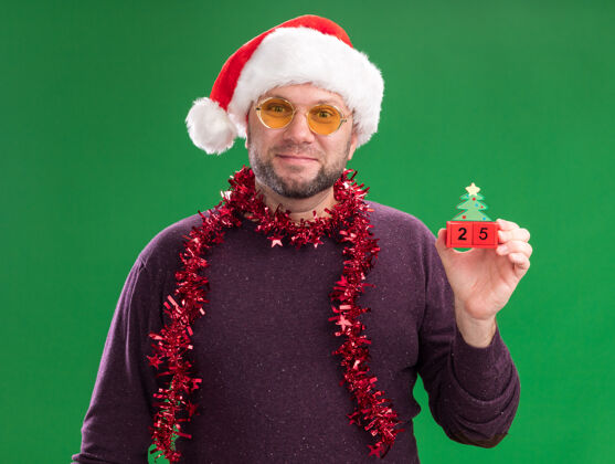 高兴高兴的中年男子戴着圣诞帽 脖子上戴着金箔花环 戴着眼镜 手里拿着圣诞树玩具 绿色的墙上挂着隔离日期穿着花环脖子