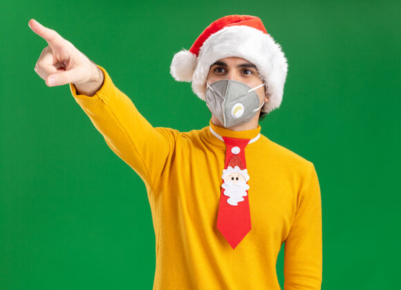 年轻人一个穿着黄色高领毛衣 戴着圣诞老人帽 戴着滑稽领带 戴着护面面具的年轻人一边看一边用食指指着绿色墙上站着的东西领带指指点点圣诞老人