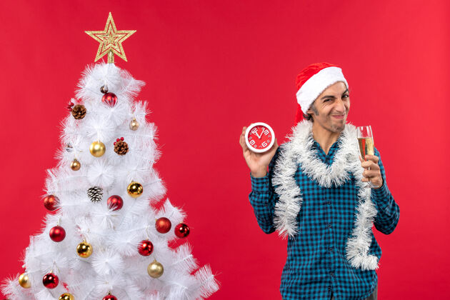葡萄酒一个戴着圣诞老人帽子 举着一杯葡萄酒 拿着一个挂钟的酸溜溜的年轻人站在红色的圣诞树旁玻璃帽子庆祝