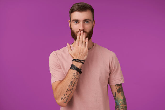 男性迷人的年轻纹身蓝眼睛男子纹身穿米色t恤和时髦的配件 而在紫色摆姿势 保持手掌放在嘴上 看起来积极衣服身体纹身