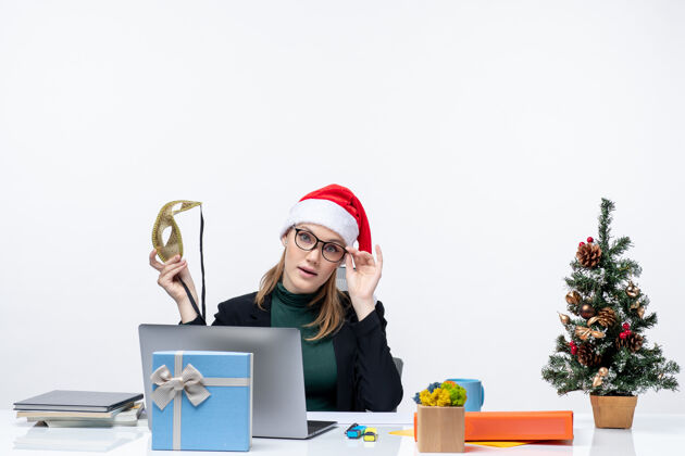 自由职业者戴着圣诞老人帽 戴着眼镜 拿着面具的年轻女子坐在桌子旁 手里拿着圣诞树和礼物工作空间树工作