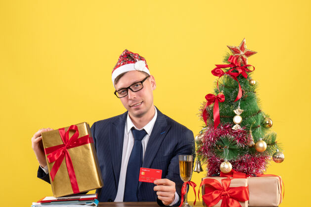 新郎商务人士拿着圣诞帽 拿着卡片和礼物坐在圣诞树旁的桌子旁 黄色的礼物商人生意人黄色