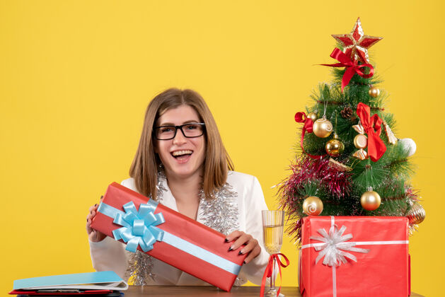 快乐正面图：女医生坐在桌子前 拿着礼物 黄色背景上的圣诞树和礼盒健康医生微笑