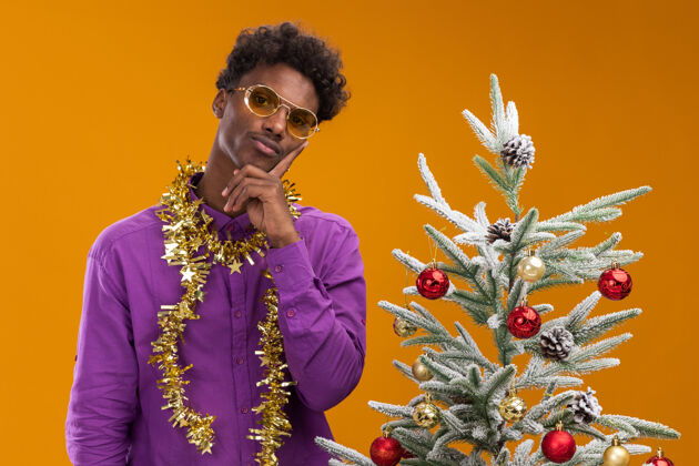 站体贴的美国黑人年轻人戴着一副眼镜 脖子上戴着金箔花环 站在橙色背景的装饰过的圣诞树旁装饰金属片近