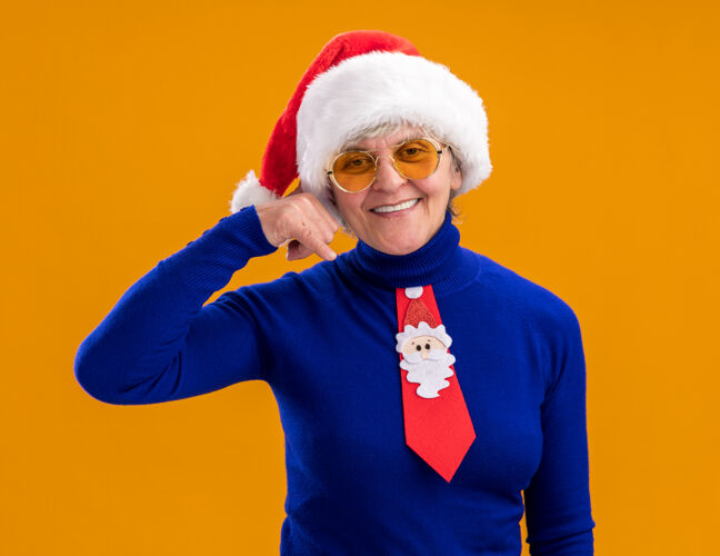 帽子戴着太阳眼镜 戴着圣诞帽 系着圣诞领带 微笑着的老妇人 打着“呼叫我”的手势 橙色背景上有一个单独的复制空间背景圣诞老人年
