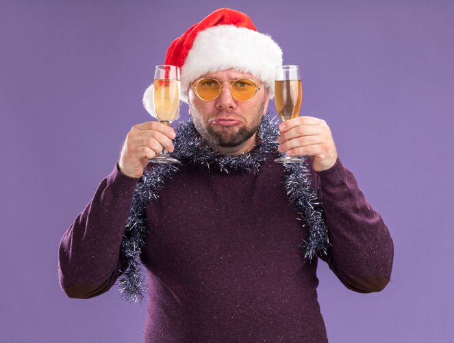 周围悲伤的中年男子戴着圣诞帽 脖子上戴着金箔花环 戴着一副眼镜 手里拿着两杯香槟 隔离在紫色的墙上花环悲伤香槟