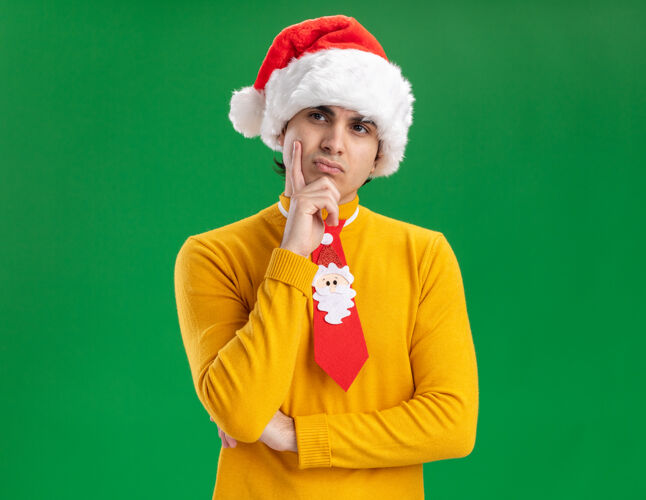 年轻穿着黄色高领毛衣 戴着圣诞老人帽 打着滑稽领带 手放在下巴上 神情沉思 站在绿色背景上思考帽子搞笑表情