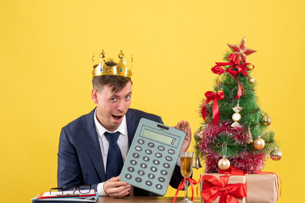 商务员商务人士手持计算器坐在圣诞树旁的桌子前 呈现黄色计算器键盘乐器人