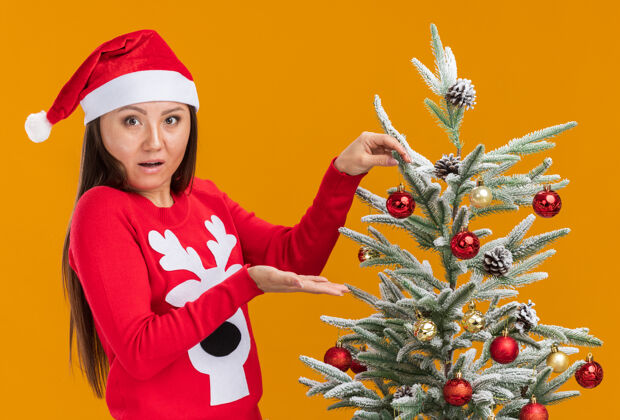 装饰惊讶的年轻亚洲女孩戴着圣诞帽和毛衣装饰圣诞树隔离在橙色的墙上圣诞节毛衣年轻