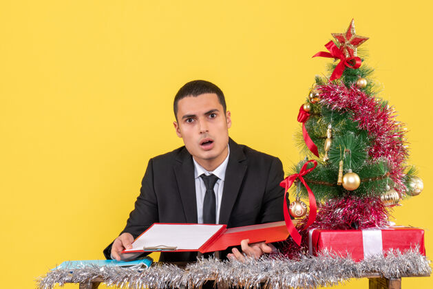 坐着前视图困惑的西服男子坐在桌子旁拿着文件圣诞树和礼物男人在西装圣诞笔记本电脑