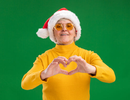 圣诞老人微笑的老妇人戴着太阳眼镜 戴着圣诞老人的帽子 在绿色的背景上用复制的空间做着孤立的心的手势老人手势眼镜