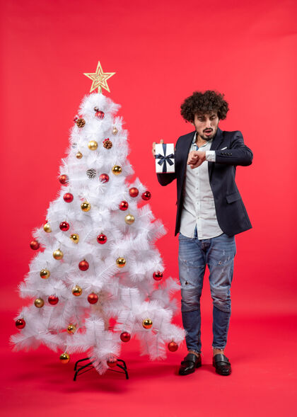 礼物年轻人在装饰的白色圣诞树旁展示他的礼物和检查时间庆祝新年男人年轻人圣诞节
