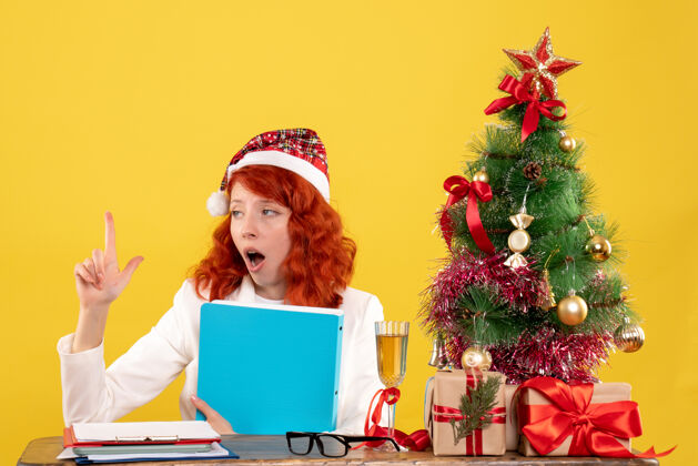 坐前视图：女医生坐在桌子后面 手里拿着文件 背景是黄色的圣诞树和礼盒医生圣诞节健康