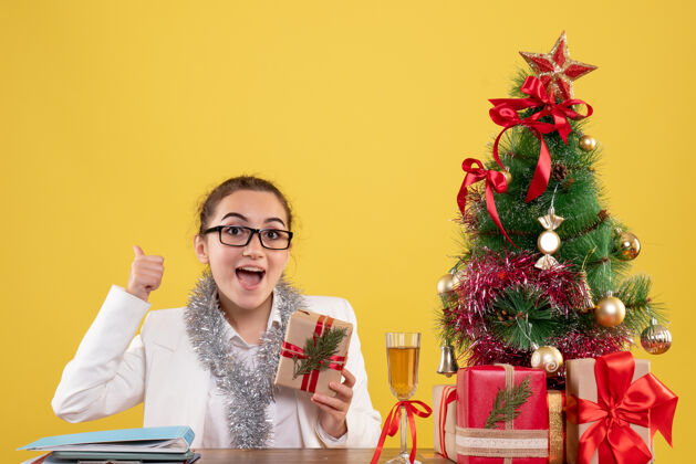 插花前视图：女医生拿着圣诞礼物和黄色背景上的圣诞树坐着女医生快乐人