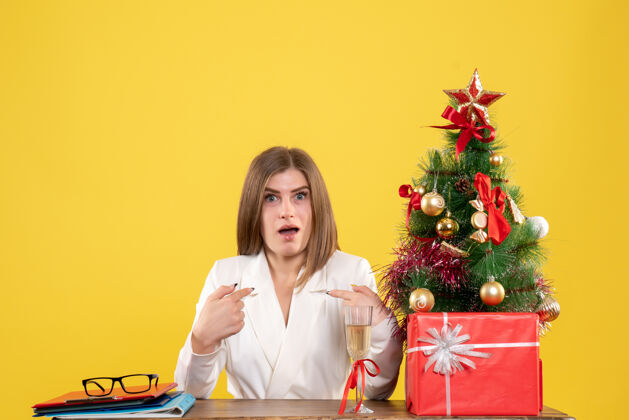 快乐前视图女医生坐在桌子前面 黄色背景上有圣诞礼物和圣诞树安排树礼物