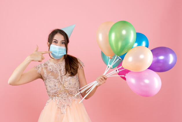 举行戴着派对帽和医用面具的快乐女孩指着她的面具 手里拿着粉色的彩色气球粉红色乐趣面具