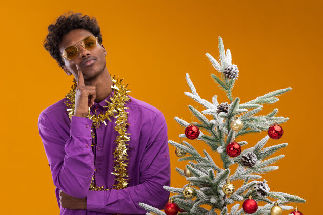 围着自信的非洲裔美国年轻人戴着眼镜 脖子上戴着金箔花环 站在装饰过的圣诞树旁 抚摸着隔离在橙色墙上的脸站着非裔美国人年轻人
